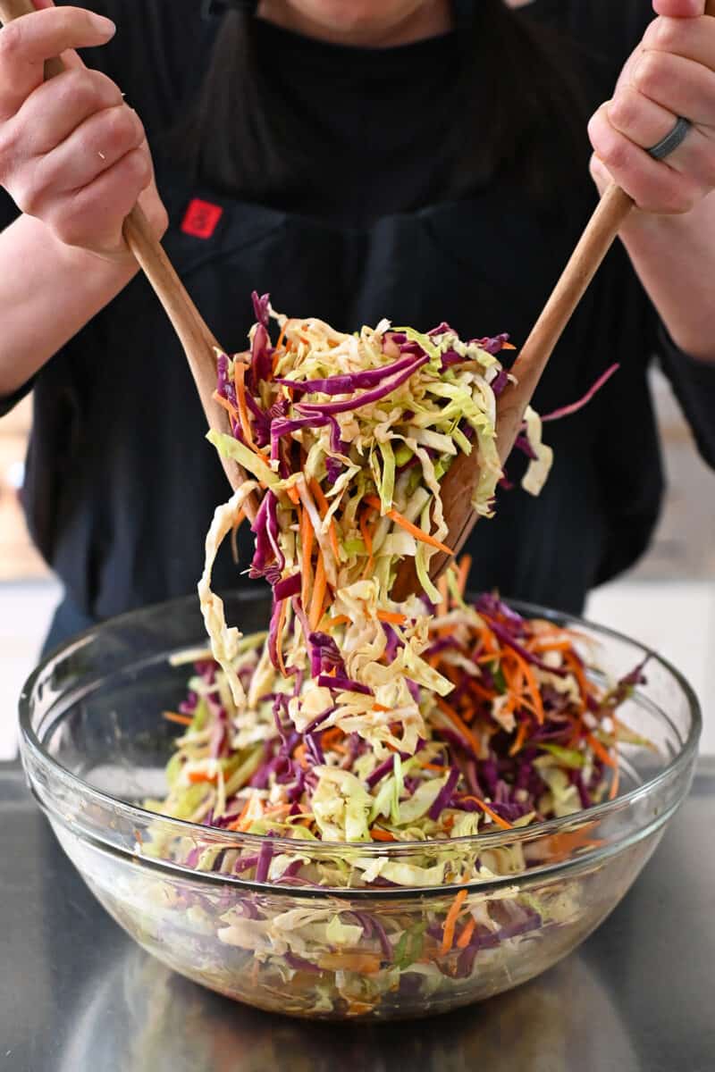 A person in a black apron is tossing pineapple coleslaw in a large clear bowl.