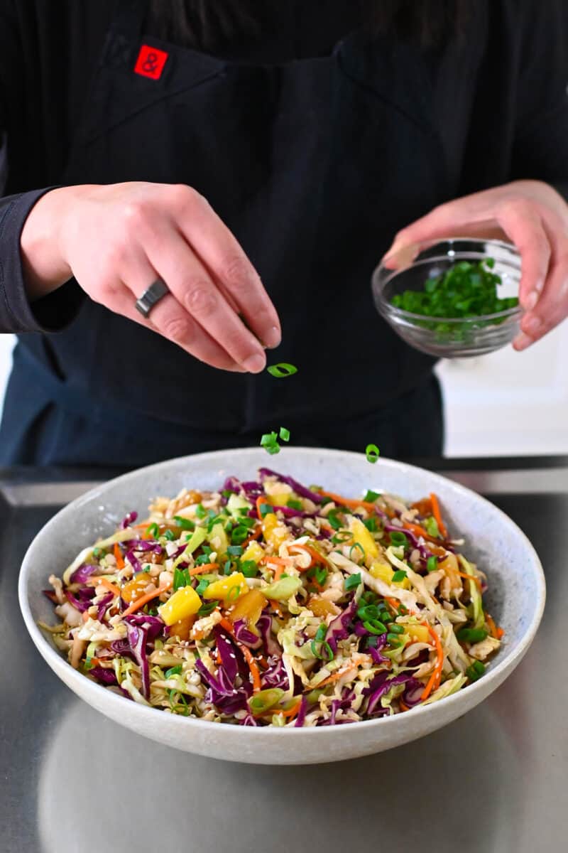 A hand is adding sliced green onions to a platter filled with pineapple coleslaw.