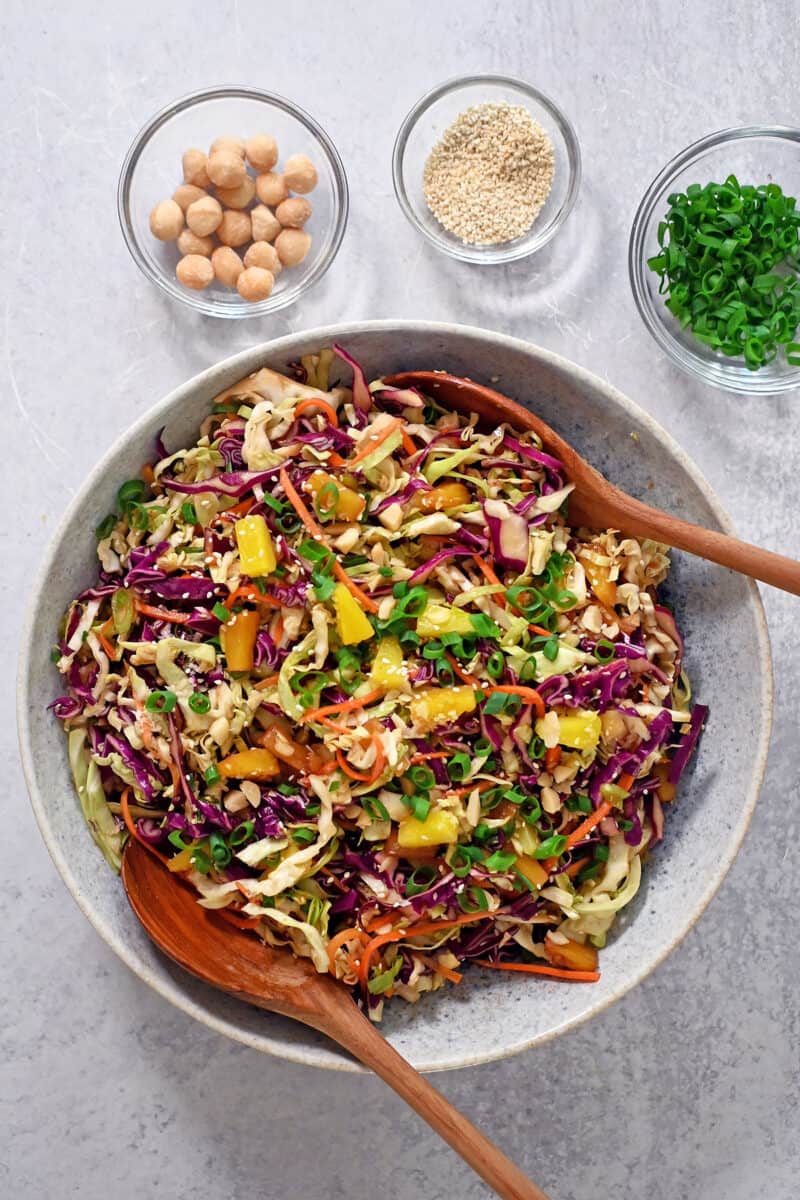 An overhead shot of pineapple coleslaw next to small bowls of macadamia nuts and toasted sesame seeds.
