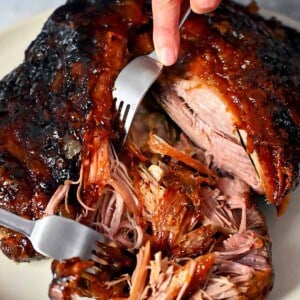 Two forks are shredding a pork shoulder roast that is topped with a peach barbecue sauce.