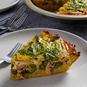A slice of paleo and dairy free asparagus quiche on a plate in front of pie pan with quiche inside.