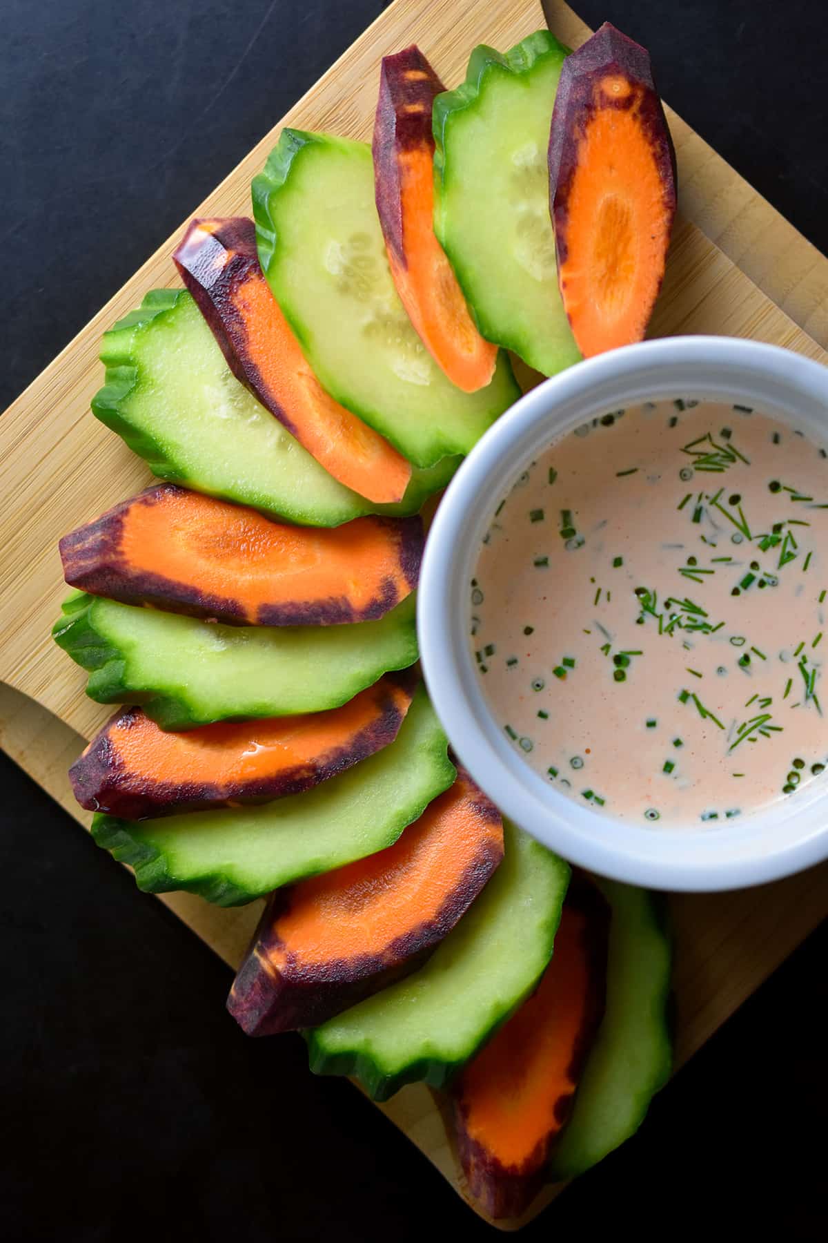 A small bowl of sriracha ranch dip is surrounded by sliced cucumber and purple carrots.