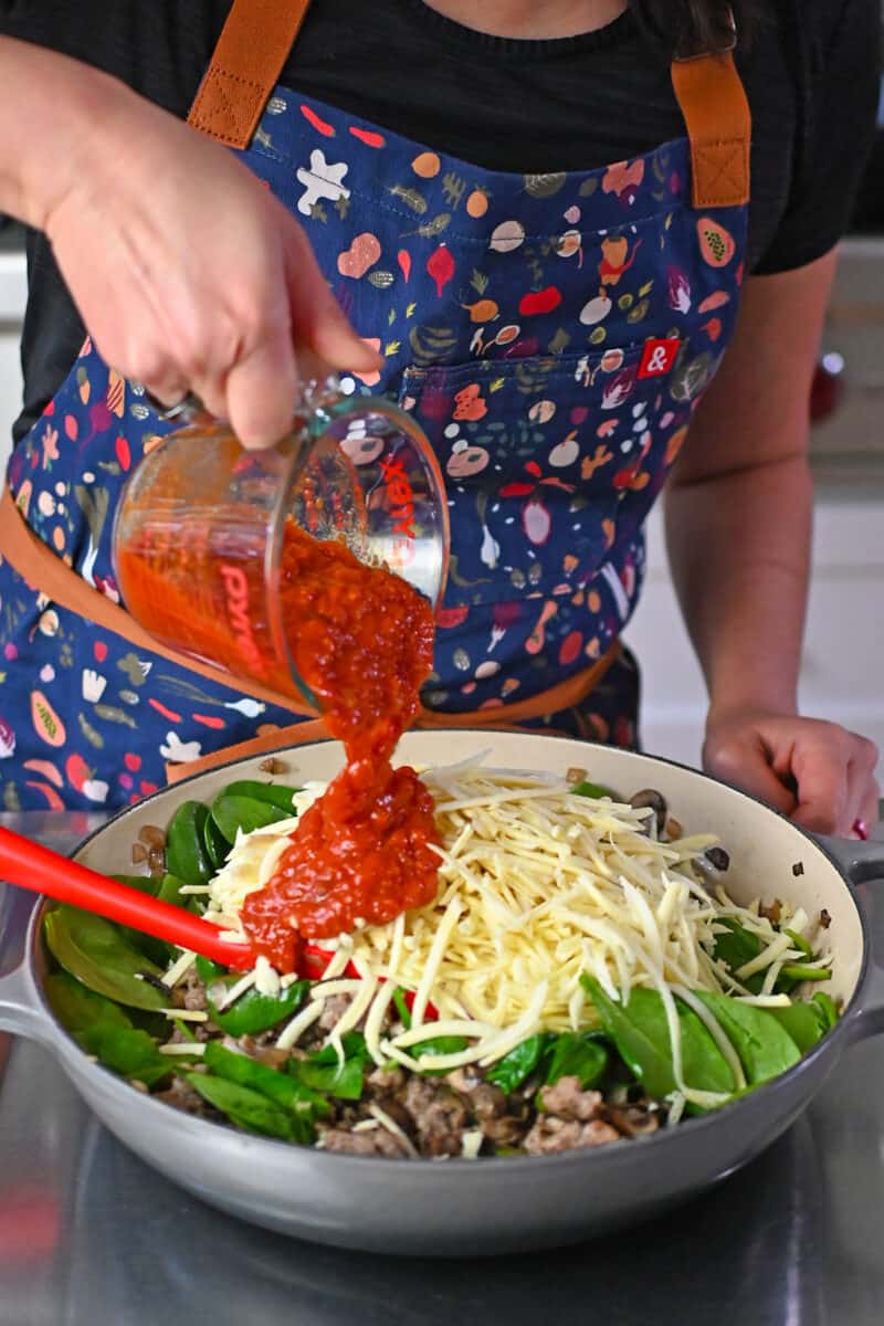 Pouring marinara sauce to a skillet filled with Italian sausage, onions, mushrooms, and shredded white sweet potatoes.
