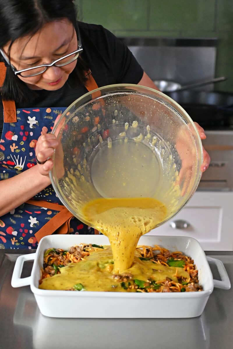 Pouring seasoned whisked raw eggs into a casserole pan filled with sausage and vegetables.