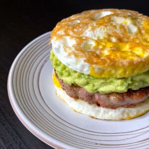 A white plate topped with a paleo egg McMuffin breakfast sandwich made with two fried egg buns and a sausage patty filling and guacamole.