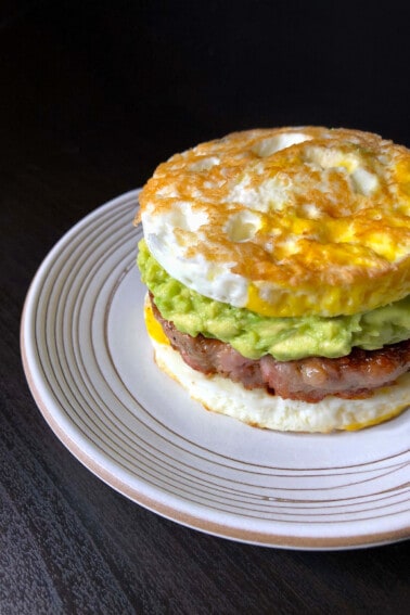 A white plate topped with a paleo egg McMuffin breakfast sandwich made with two fried egg buns and a sausage patty filling and guacamole.