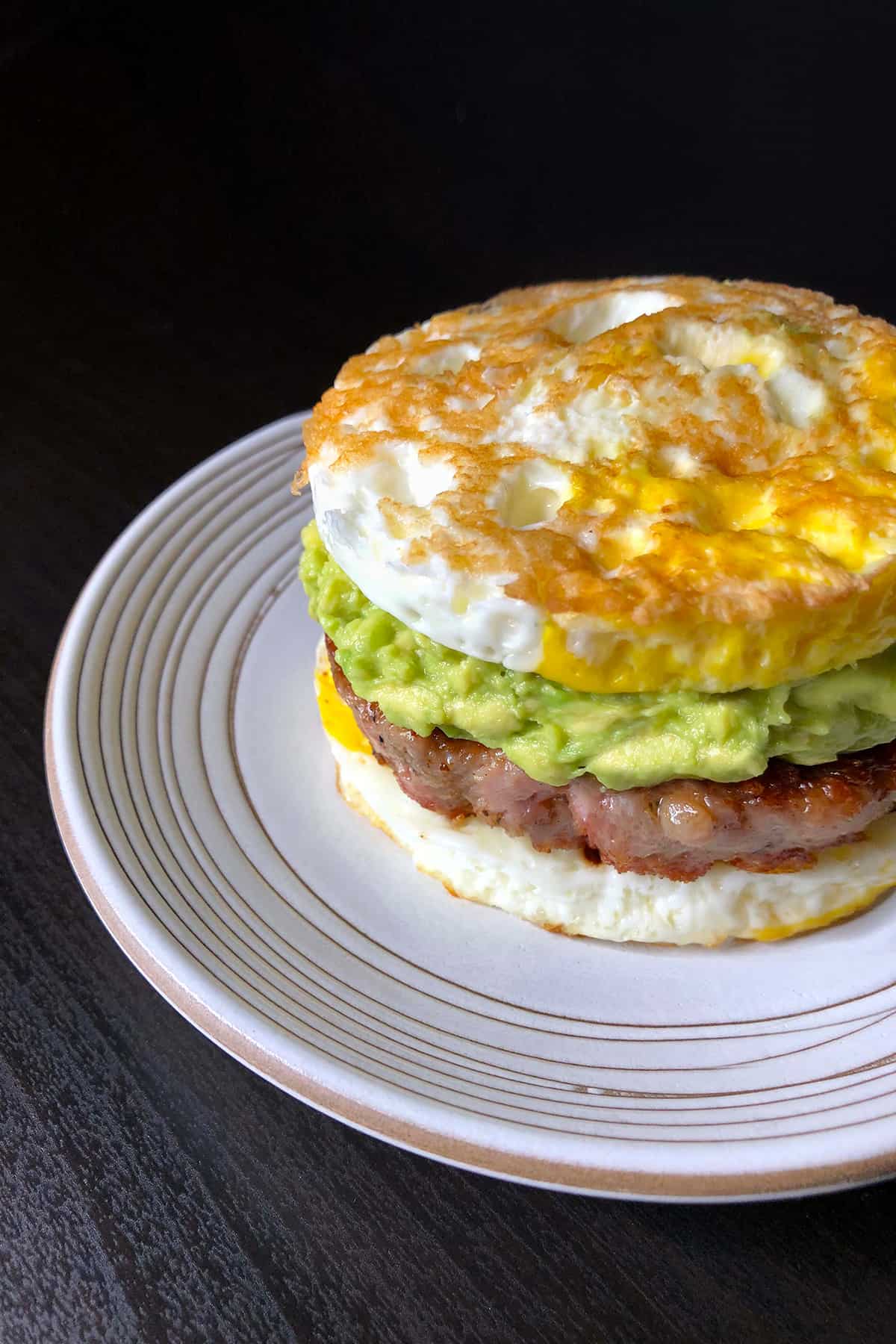 A white plate topped with a paleo egg McMuffin breakfast sandwich made with two fried egg buns and a sausage patty filling and guacamole.
