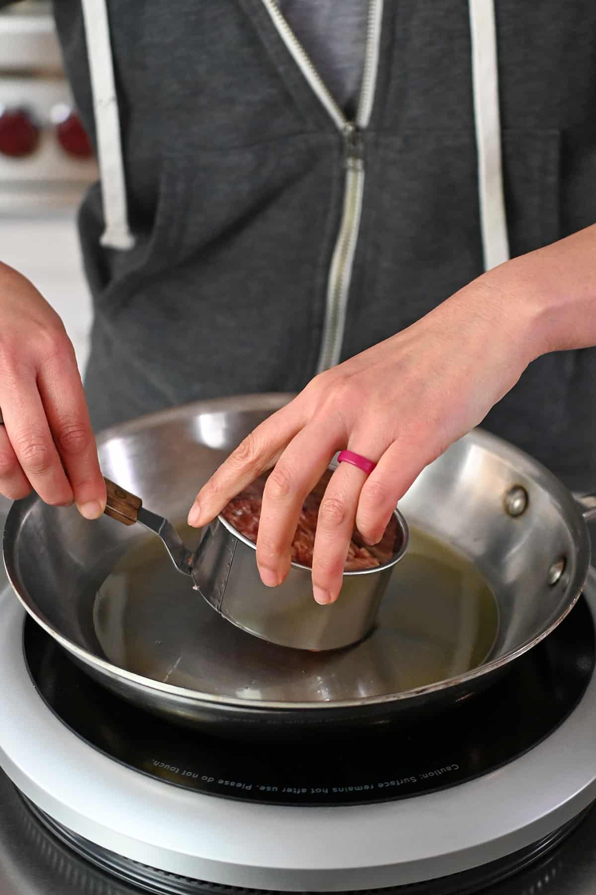 Adding a biscuit cutter filled with raw sausage into a frying pan.