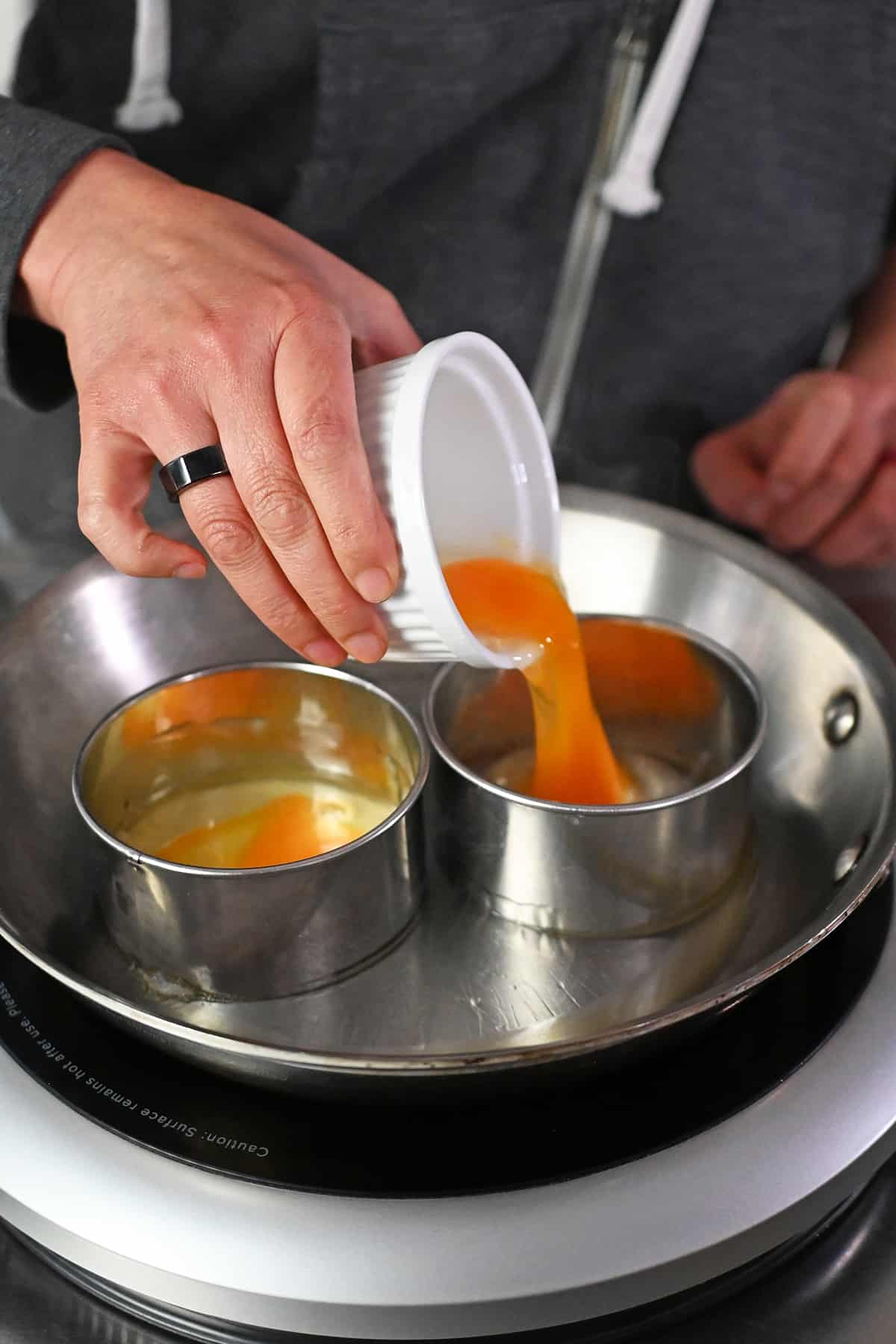 Pouring a raw egg from a white ramekin into a biscuit cutter in a frying pan.
