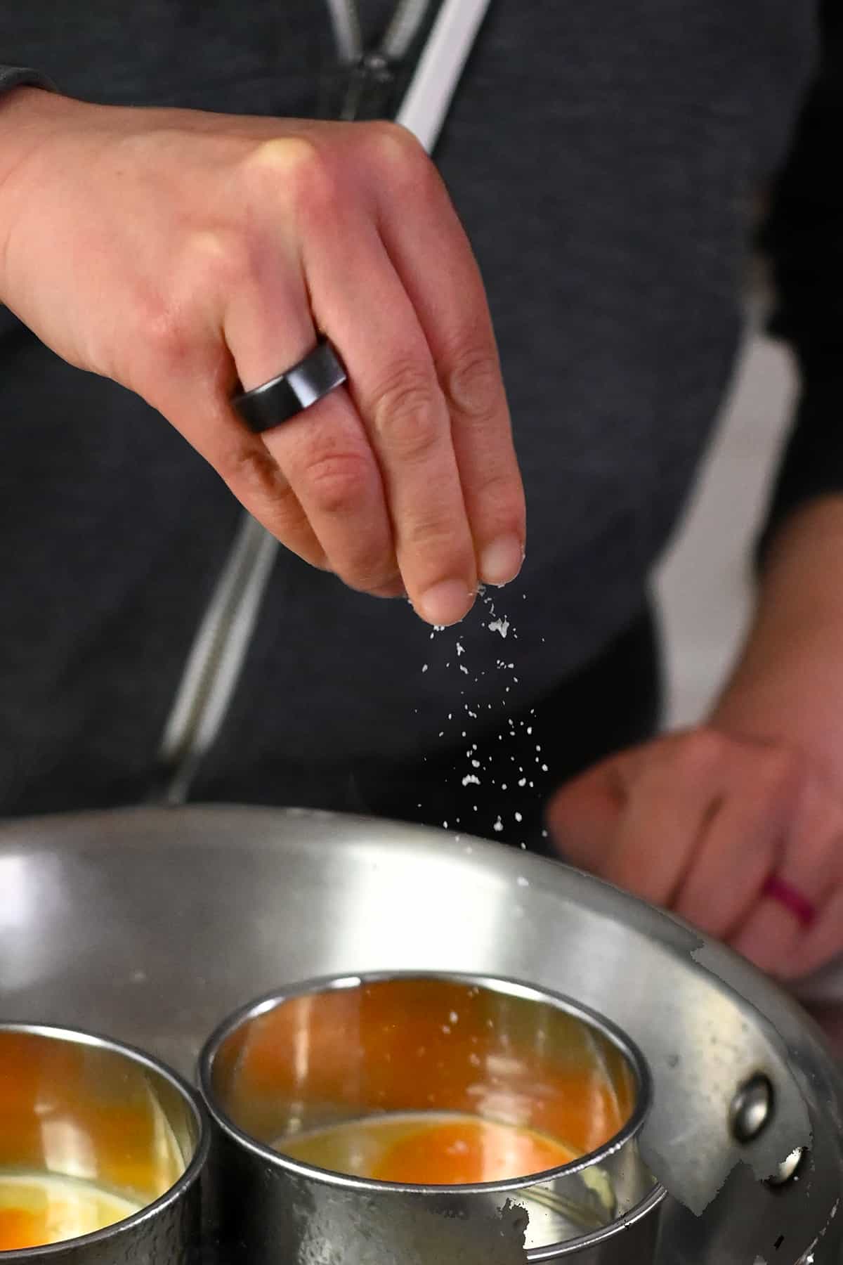 Sprinkling salt on eggs frying in a biscuit cutter in a frying pan.