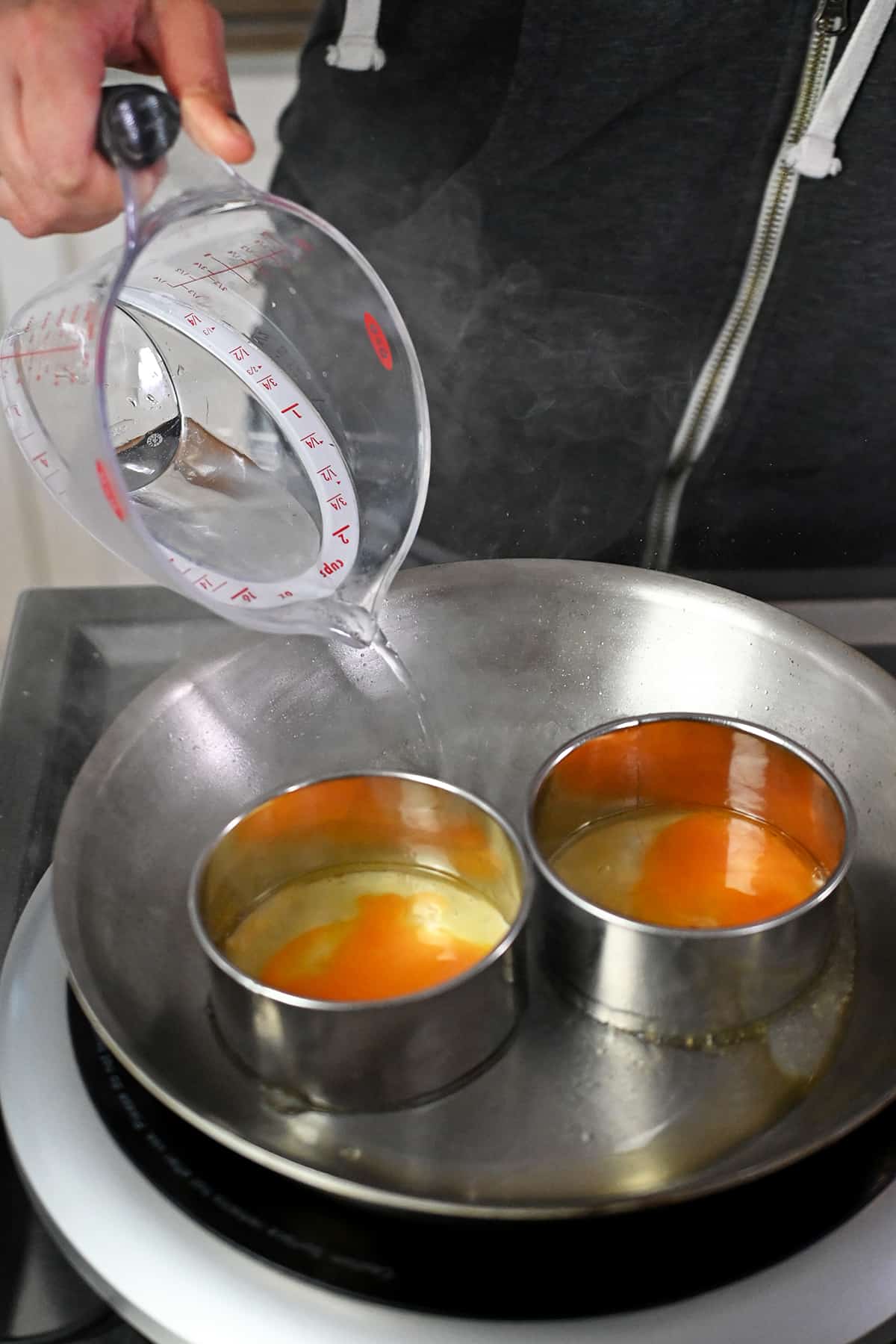 Pouring water into a skillet with two biscuit cutters filled with eggs.