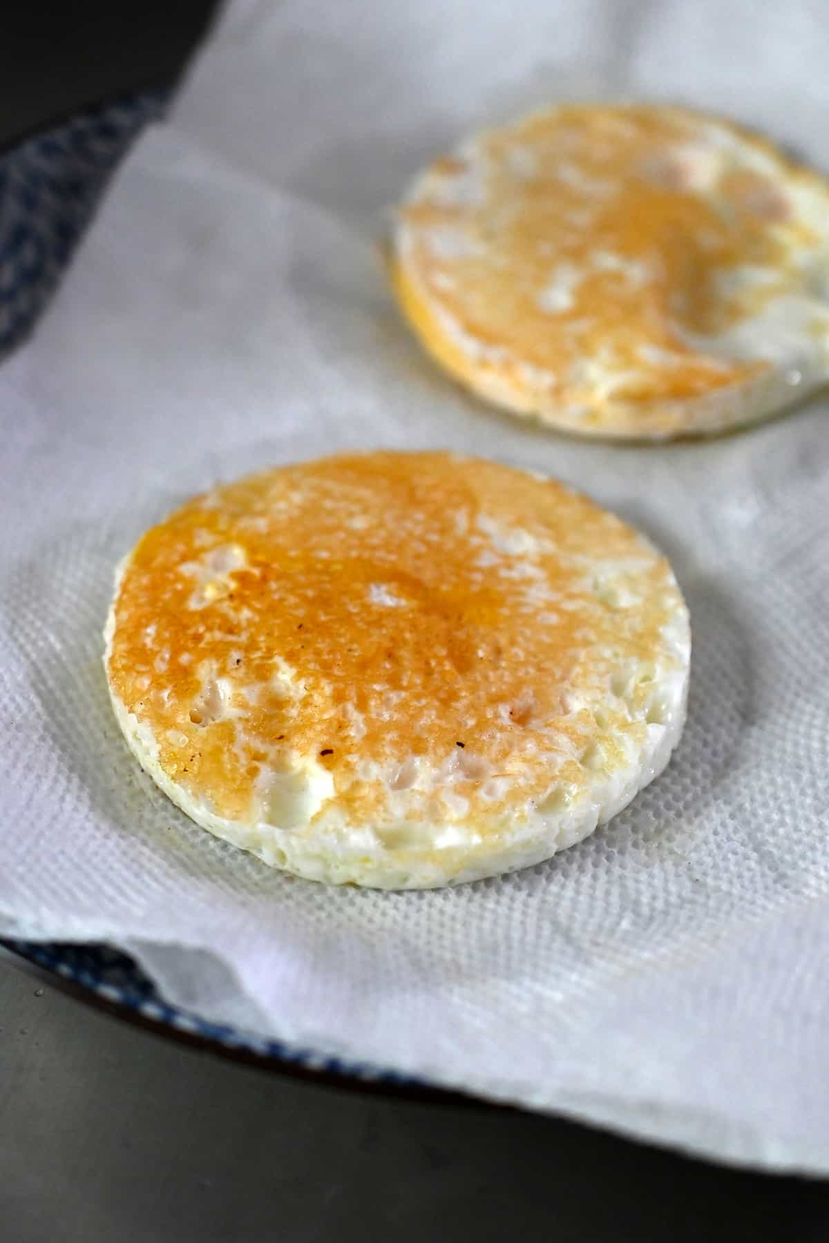 Two round fried eggs are on a paper towel lined plate.