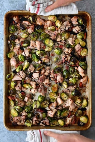 An overhead shot of two hands holding a sheet pan filled with chicken, Brussels sprouts, and bacon.