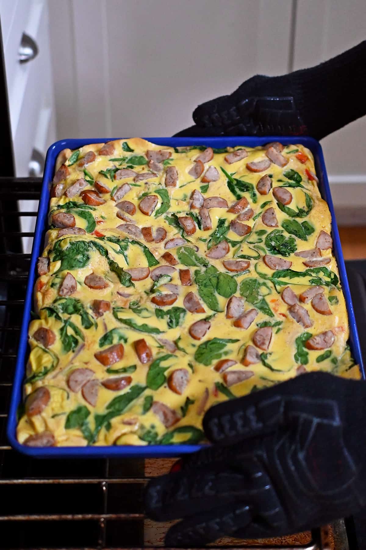Removing a freshly baked sheet pan filled with scrambled eggs, veggies, and sliced chicken apple sausage out of an oven.