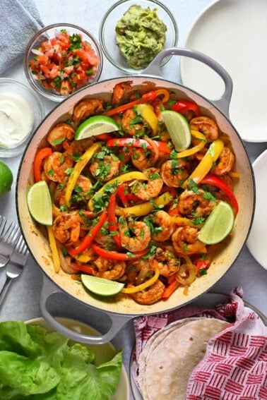An overhead shot of a skillet filled with shrimp fajitas surrounded by bowls filled with guacamole, sour cream, and salsa, fresh lettuce leaves, and tortillas.