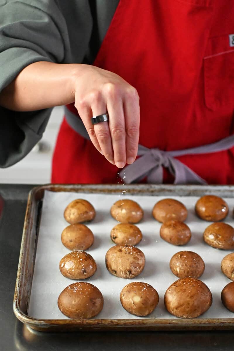 Sprinkling salt on top of ghee coated brown mushrooms on a parchment paper lined rimmed baking sheet. 