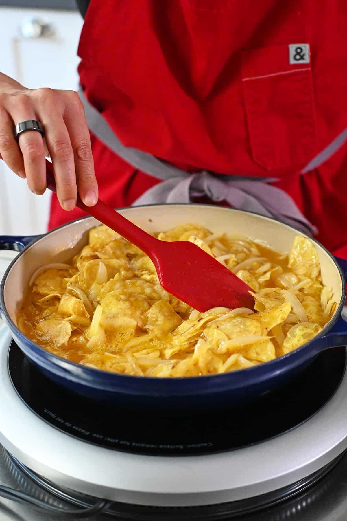 A red silicone spatula is rearranging potato chips and whisked raw eggs in a blue skillet so the top is a smooth layer.