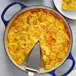 An overhead shot of a skillet filled with a Spanish tortilla made with potato chips with a missing slice that is on a plate next to the pan.