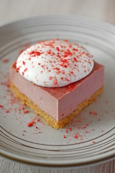 A slice of paleo, vegan, and gluten free strawberry cheesecake bars on a white plate, topped with whipped coconut cream and strawberry powder.