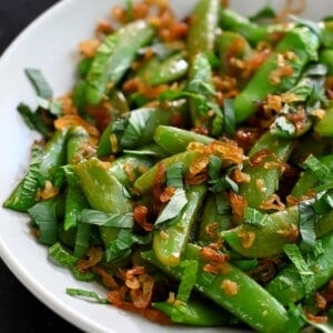 A side view of sugar snap peas sautéed with garlic, fish sauce, mint, Thai basil, and crispy shallots.