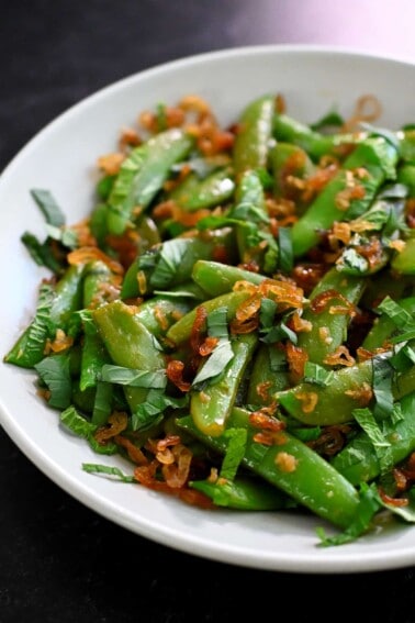 A side view of sugar snap peas sautéed with garlic, fish sauce, mint, Thai basil, and crispy shallots.