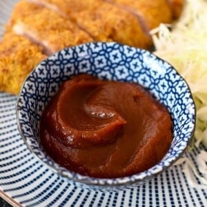 A closeup shot of a small blue bowl filled with homemade Tonkatsu Sauce next to a fried Japanese pork cutlet and shaved cabbage.