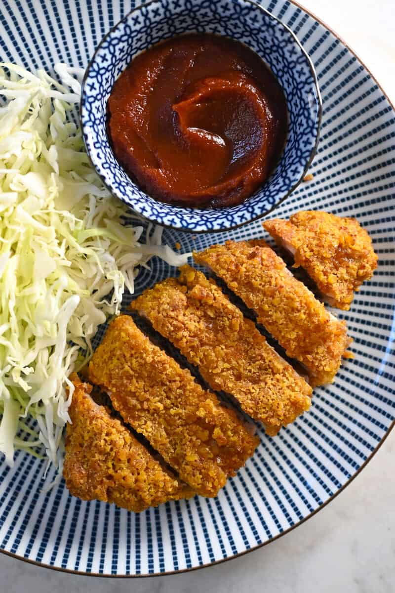 An overhead shot of a plate with paleo tonkatsu next to a pile of shredded cabbage and a small dish of homemade Tonkatsu Sauce.
