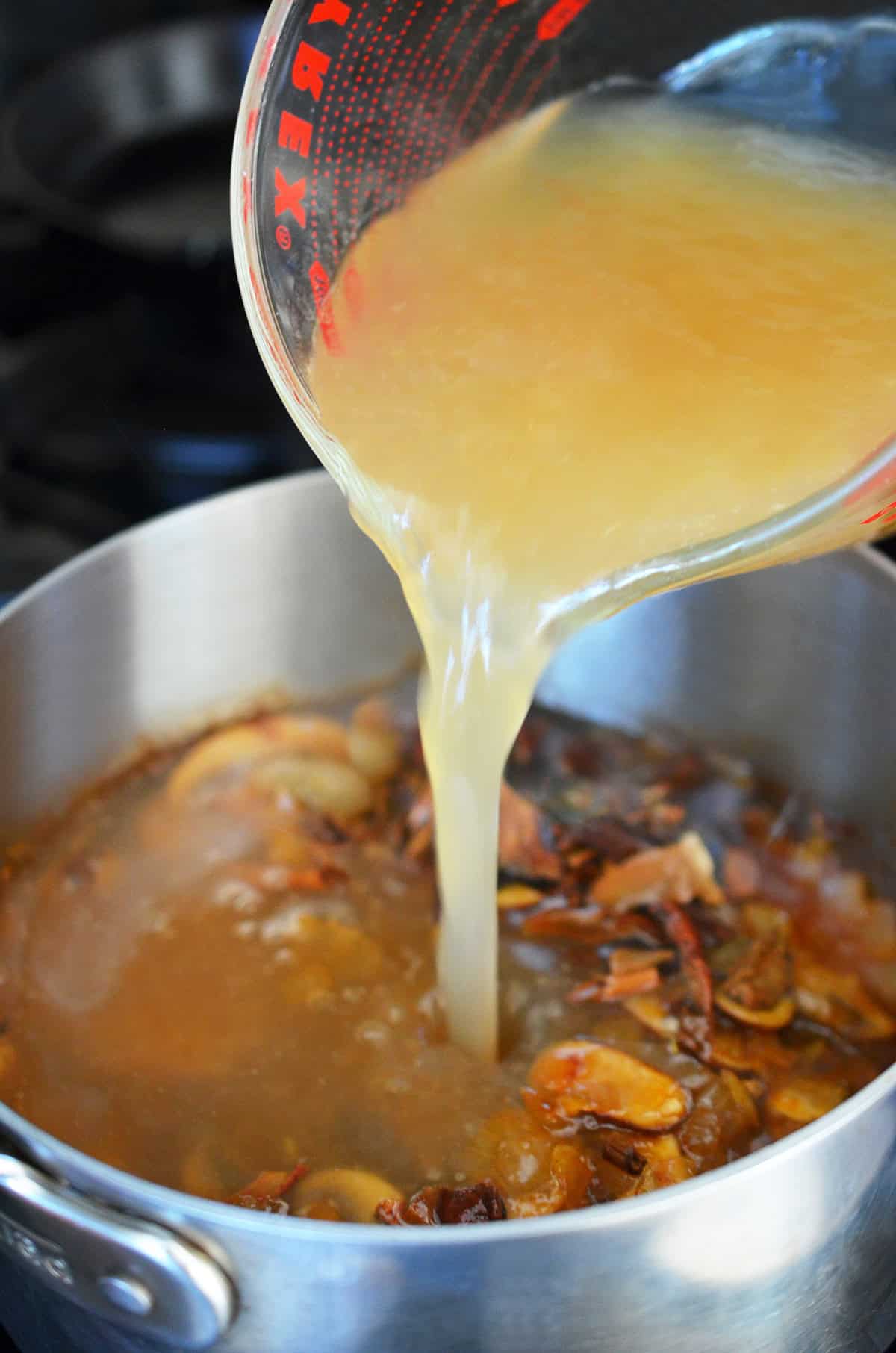 Pouring chicken broth into a saucepan filled with the ingredients for a gluten free gravy that has tons of umami flavor.