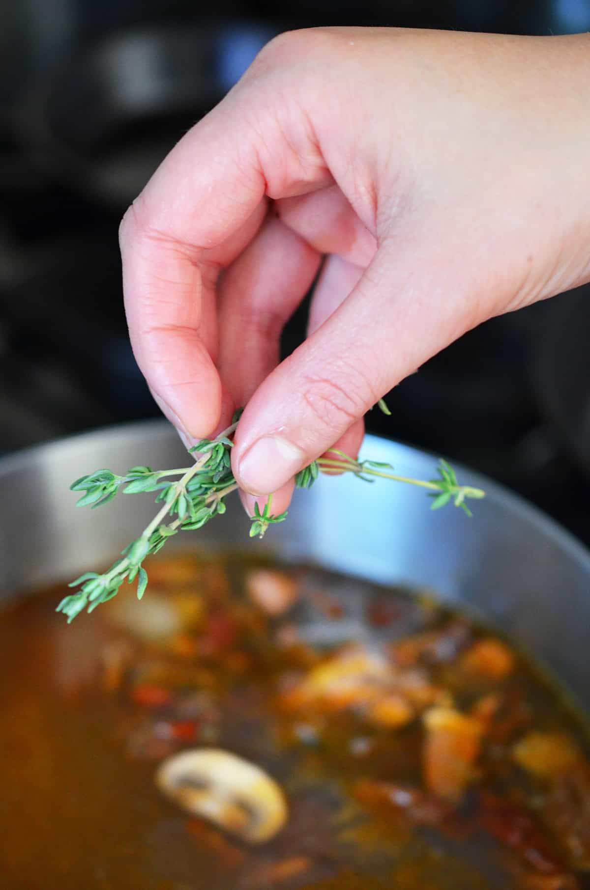 A hand is adding a fresh thyme sprig into a saucepan filled with umami gravy ingredients.