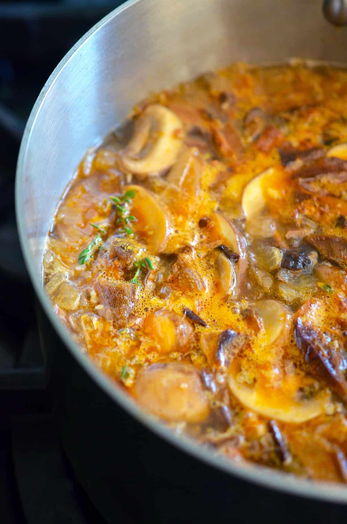 A closeup of a saucepan filled with a simmering broth filled with sliced mushrooms, thyme, onions, and broth to make umami gravy.