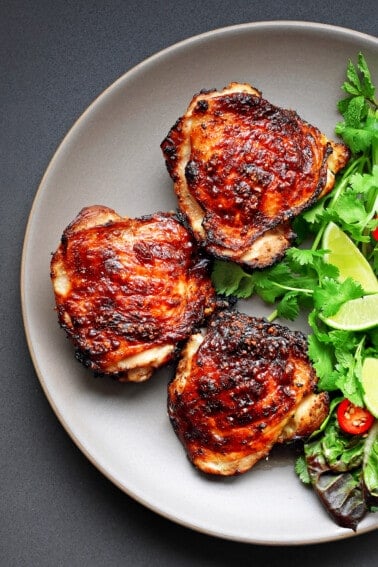 An overhead shot of a plate with three golden brown pieces of Vietnamese lemongrass chicken next to fresh herbs, lettuce leaves, chili slices, and lime wedges.