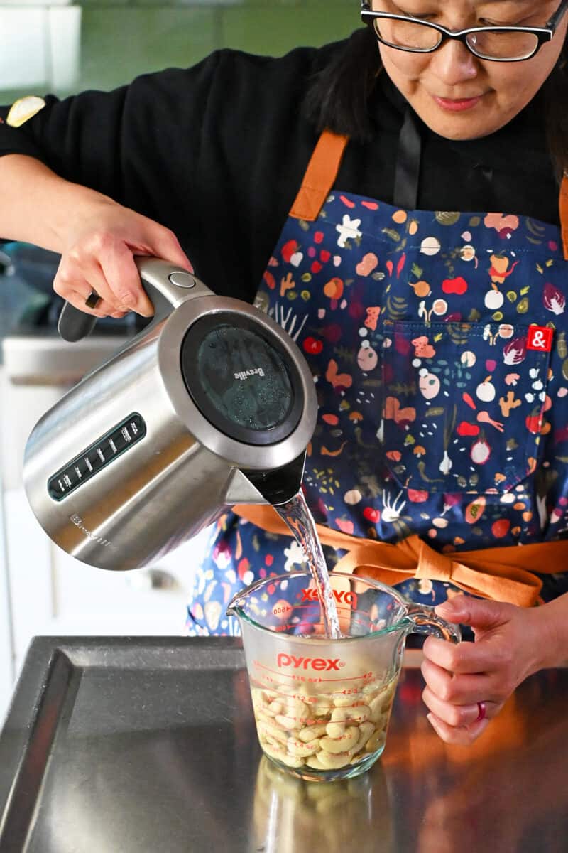 Pouring boiling water from an electric kettle into a measuring cup filled with raw cashews.
