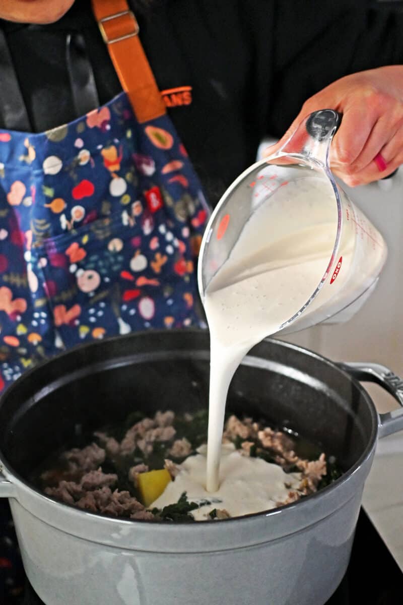 Pouring cashew cream into a pot filled with Zuppa Toscana.