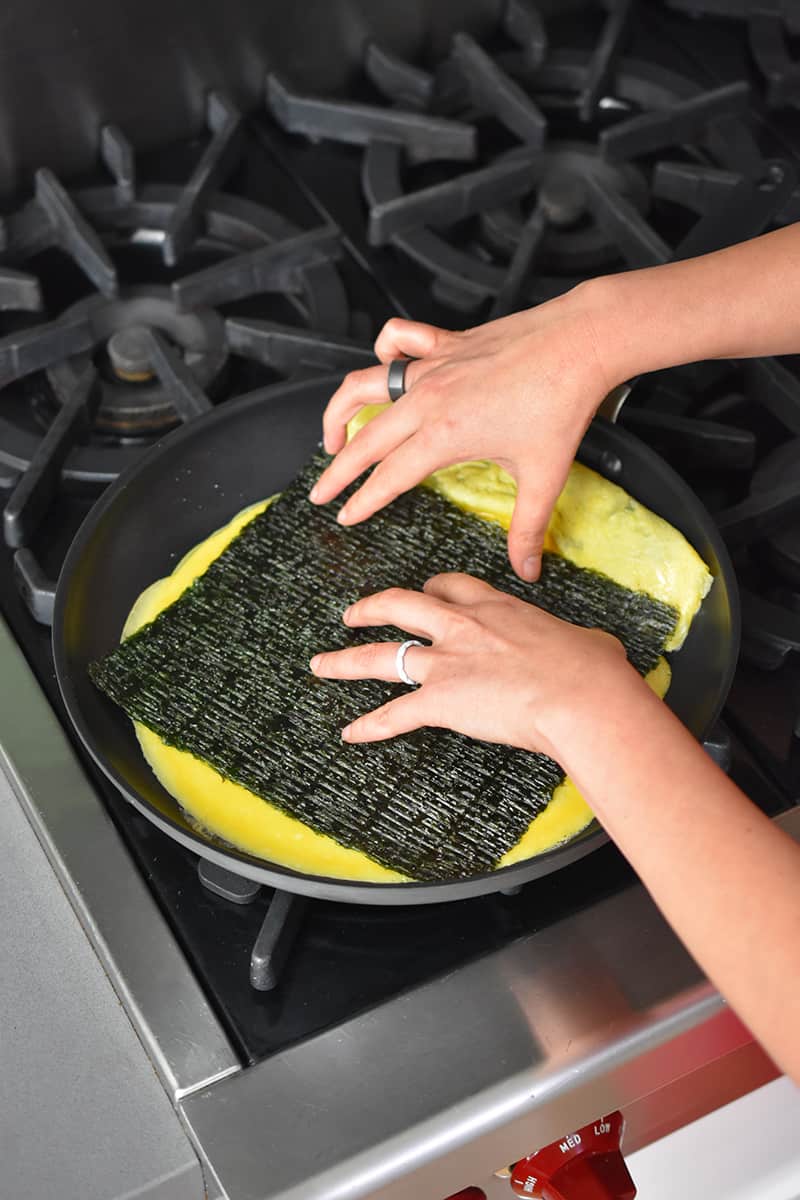 Two hands are placing a rectangular roasted seaweed in a skillet filled with a thin egg omelet.