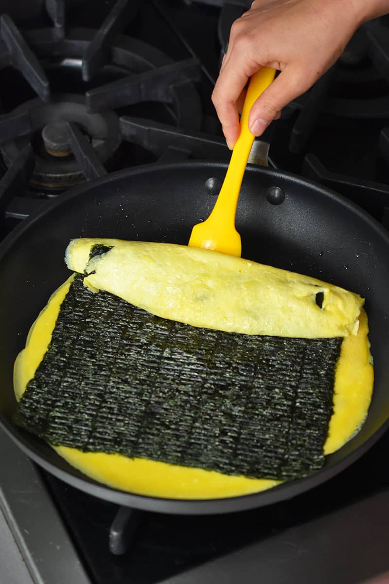 A hand is using a yellow silicone spatula to start flipping a Korean egg roll.