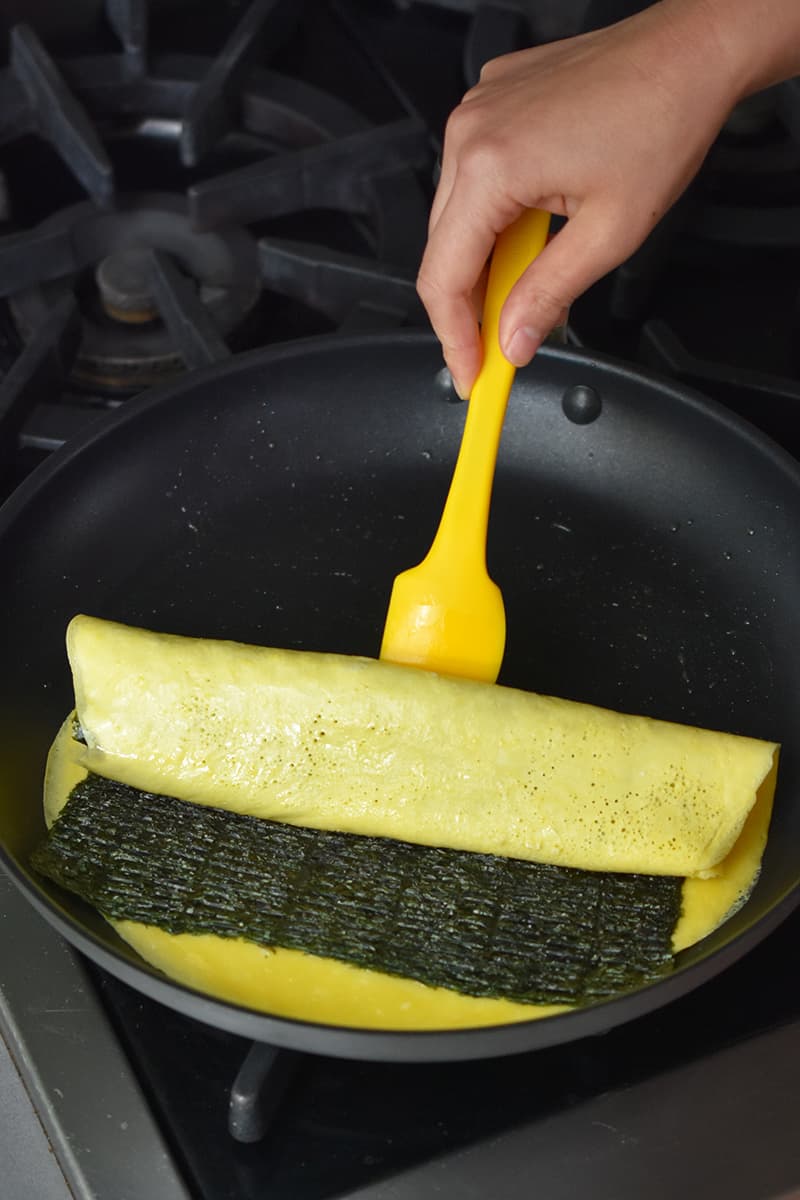 A Korean egg roll with seaweed is being rolled in a large skillet with a yellow silicone spatula.