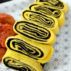 A closeup shot of a cut up Korean Egg Roll Omelet with Roasted Seaweed on a white plate with gray flowers. You can see the spirals of egg and bright yellow egg in the cut-up pieces.