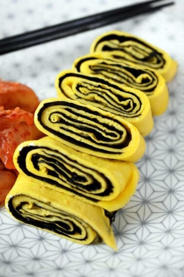 A closeup shot of a cut up Korean Egg Roll Omelet with Roasted Seaweed on a white plate with gray flowers. You can see the spirals of egg and bright yellow egg in the cut-up pieces.