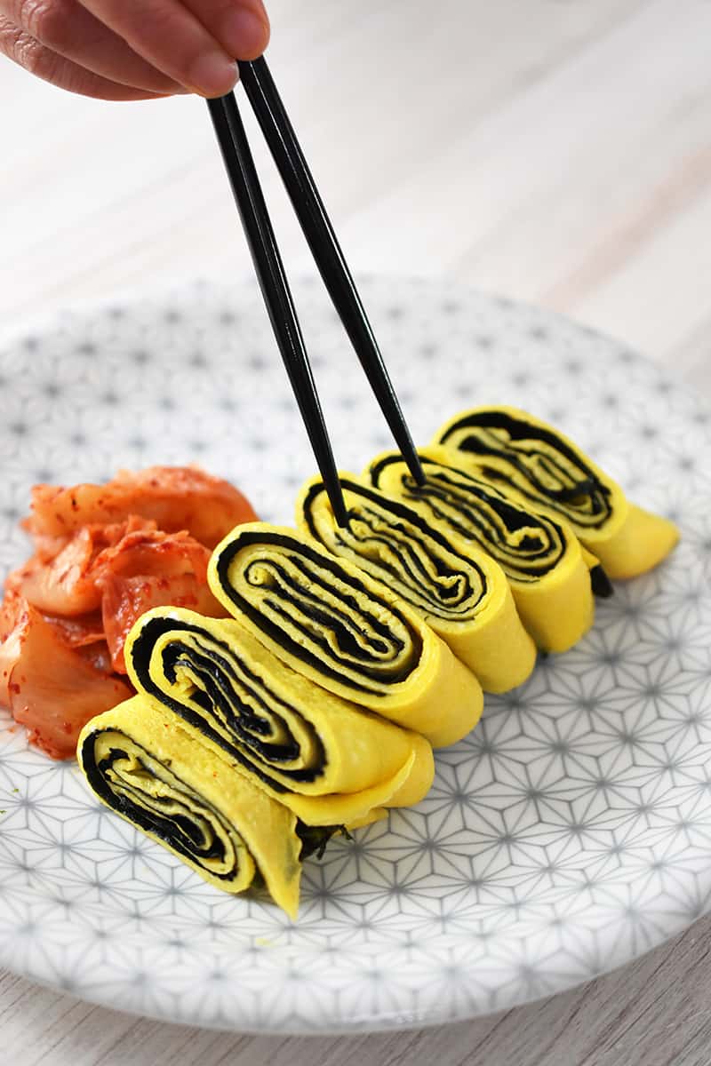 A pair of chopsticks is grabbing a slice of Korean egg roll with seaweed on a white and gray plate. There is a small pile of kimchi next to it.
