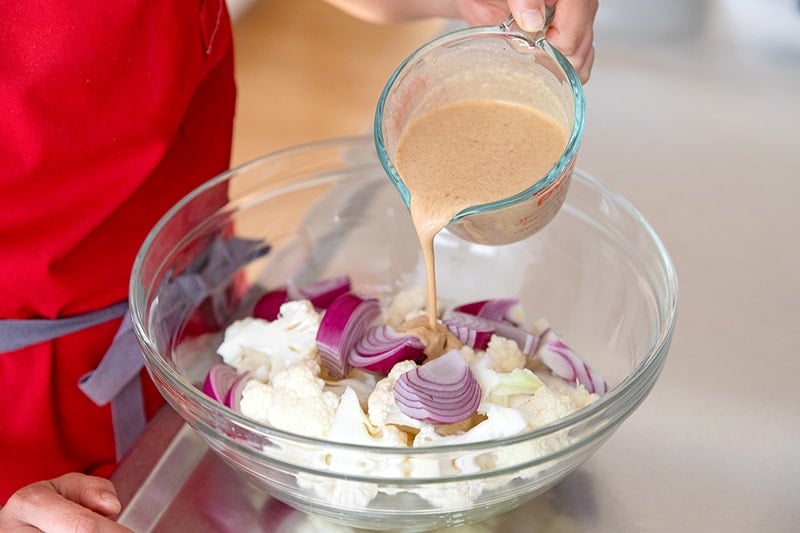 Pouring dairy-free tandoori marinade on cauliflower and onions in a large bowl for Sheet Pan Tandoori Chicken