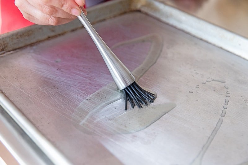 A silicone brush is spreading avocado oil on a rimmed baking sheet for a healthy sheet pan chicken dinner.