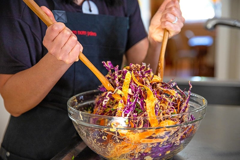 Someone tossing whole30 red cabbage slaw in a large clear bowl with wooden spoons.