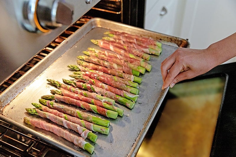 Someone placing a rimmed baking sheet of prosciutto-wrapped asparagus under the broiler in the oven.