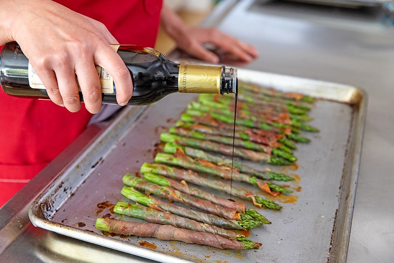 Someone adding aged balsamic to prosciutto-wrapped asparagus.