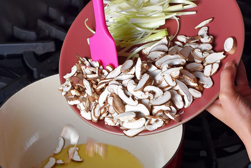 Adding sliced shiitake mushrooms and julienned leeks to a large saucepan with melted fat