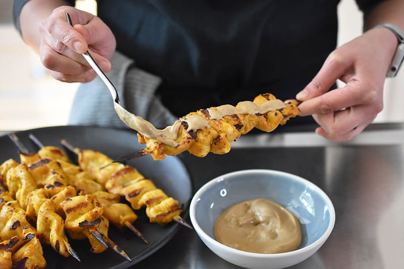 Someone spooning Hoisin No-Nut Sauce on Chicken Satay skewers.