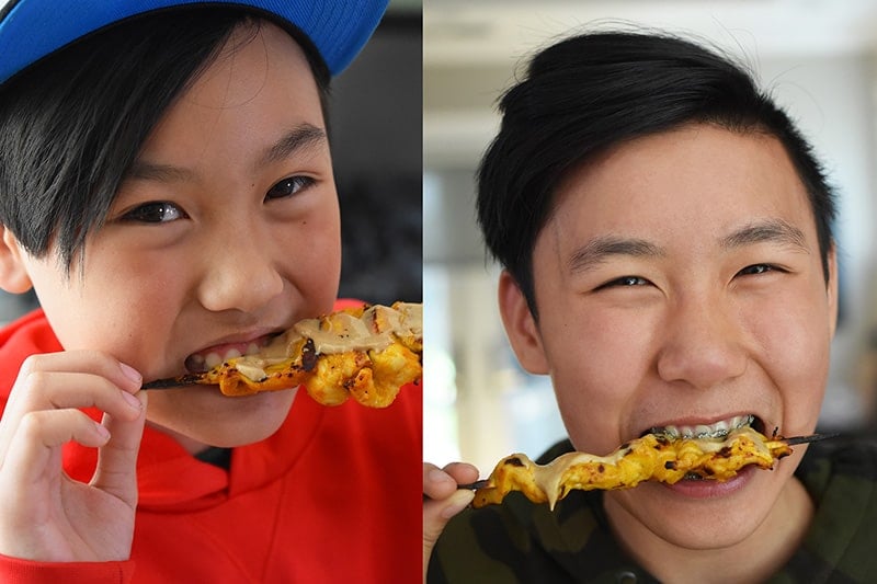 Two smiling boys biting into paleo chicken satay skewers.