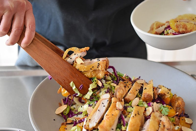 Using a pair of wooden tongs to transfer the paleo and Whole30-friendly Chinese Chicken Salad to a serving bowl. The best gluten-free Chinese Chicken Salad!