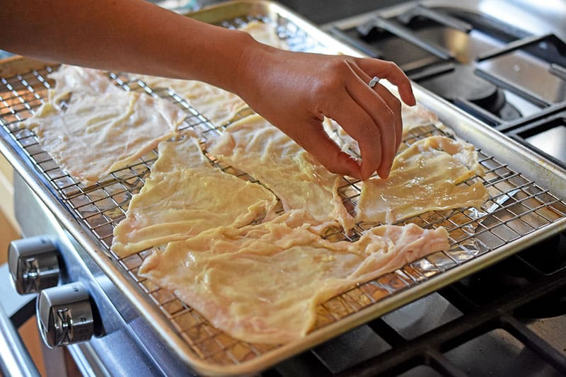 A hand placing raw chicken skins on a wire rack in a rimmed baking sheet to make oven-baked crispy chicken skins.