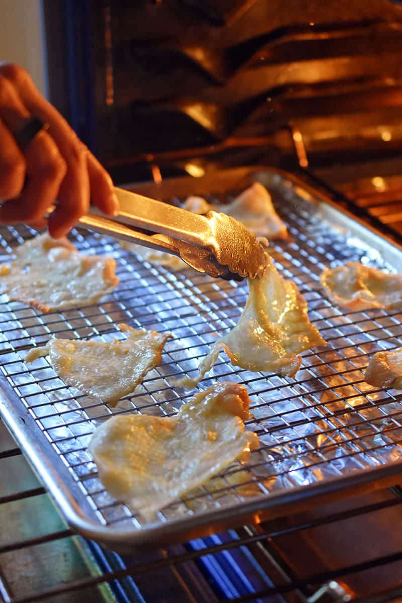 A person is using a pair of tongs to flip over chicken skins that are baking in the oven.